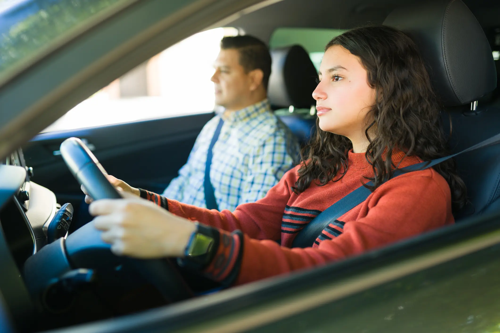 Hispanic teen girl learning to drive with her father next to her, concentrated on the road